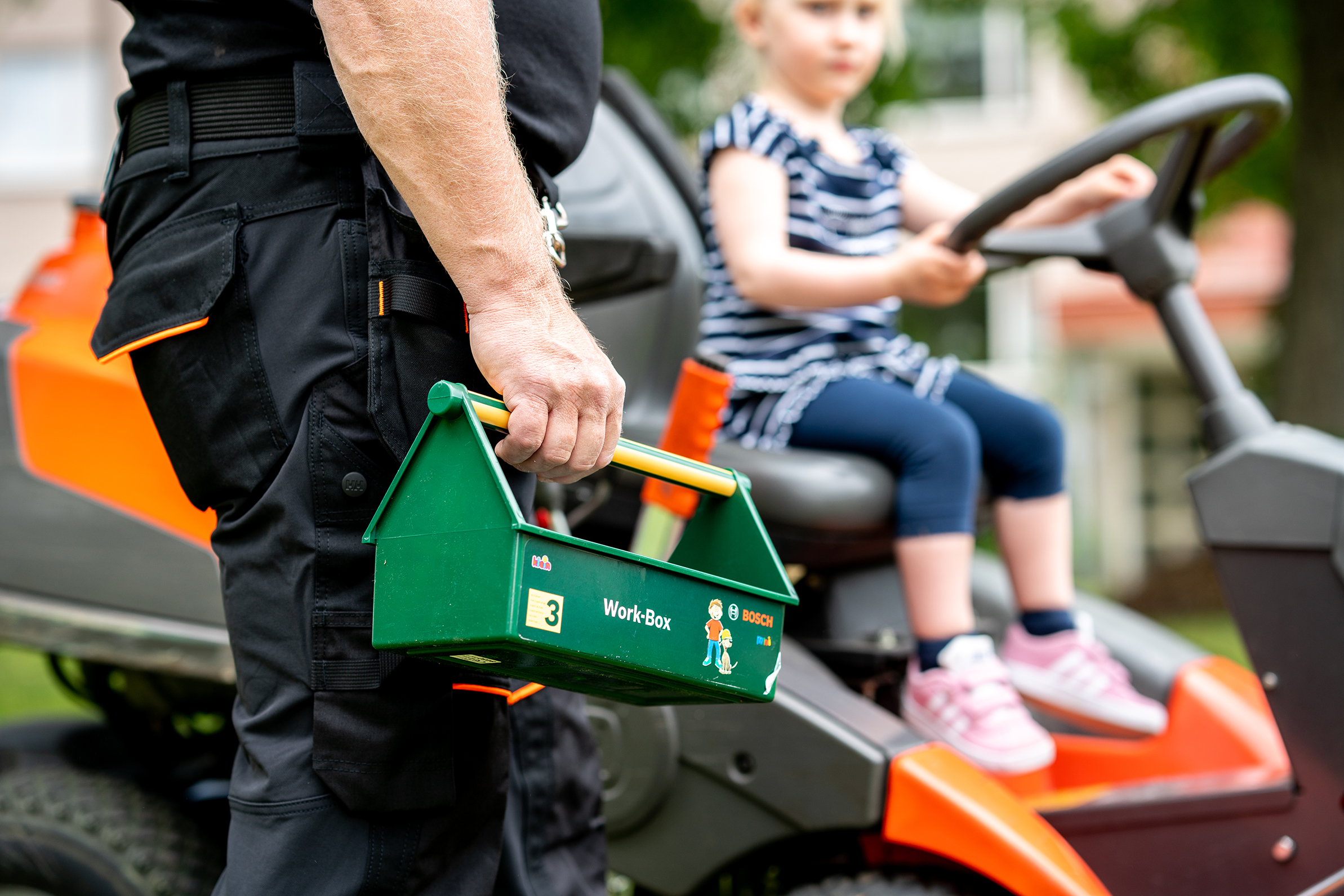 Fastighetsskötare med verktygslåda och barn på gräsklippare